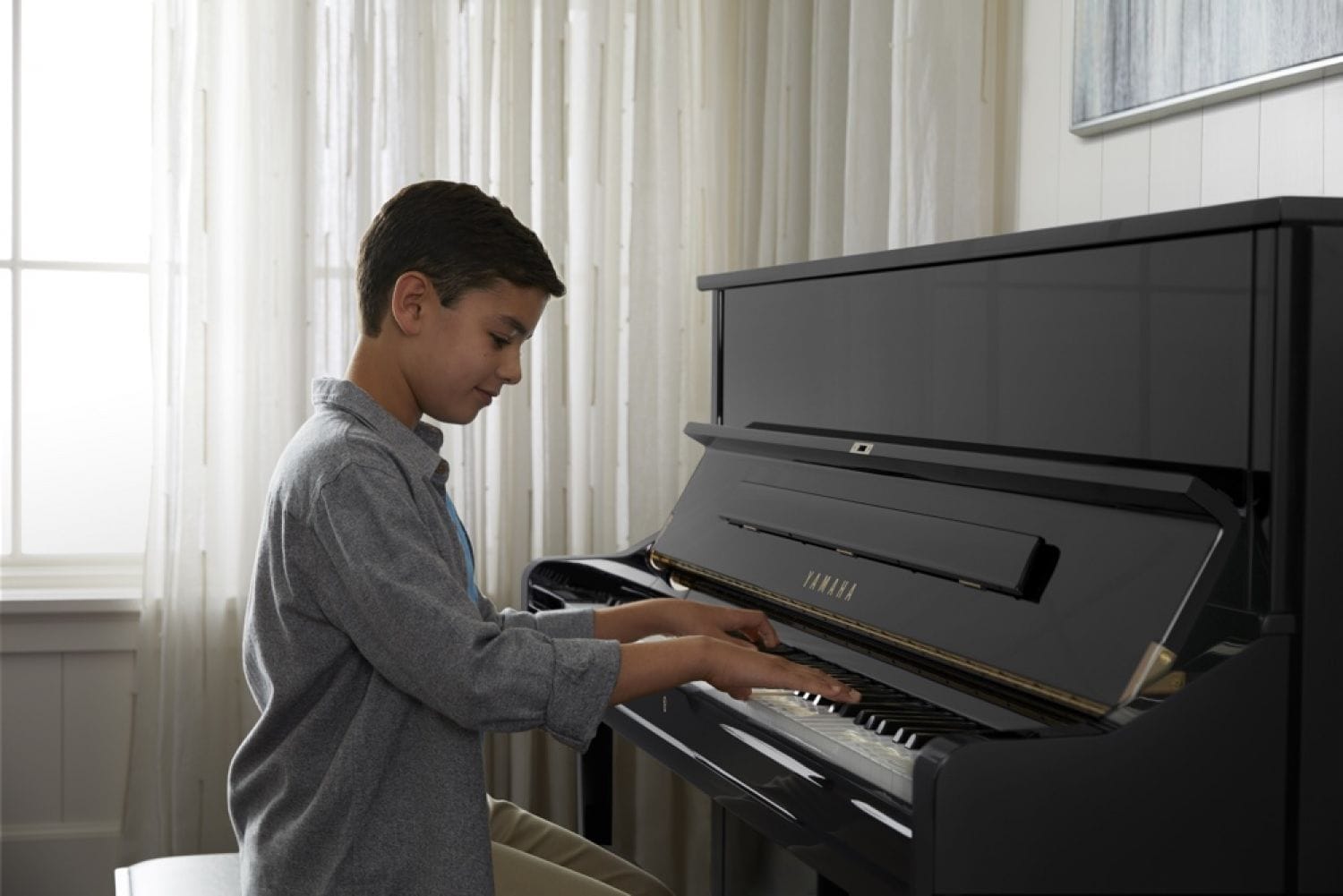 Young boy smiling as he plays a Yamaha upright piano in a living room setting.