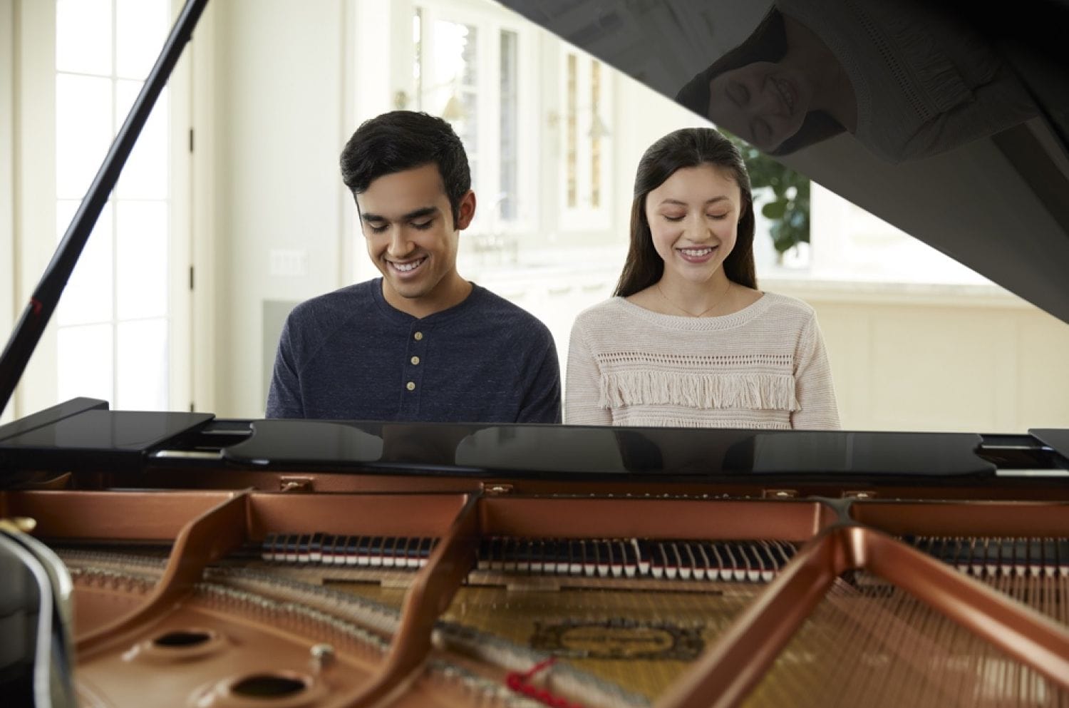 Young man and young woman seated side by side smiling and playing piano with lid open in foreground.