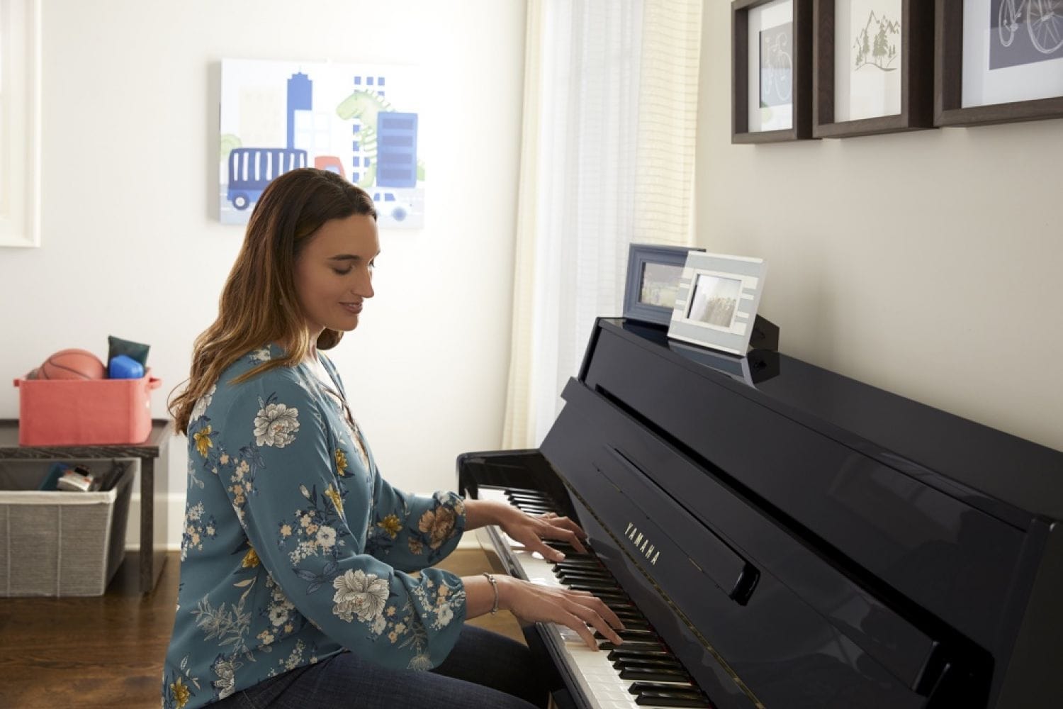 A woman approximately 30 years old with long hair and dressed casually smiling while she plays an upright piano with a lower profile back in what appears to be a kids' playroom in a home. There are kids drawings and toys in baskets in background and family pictures framed sitting on the back of piano.